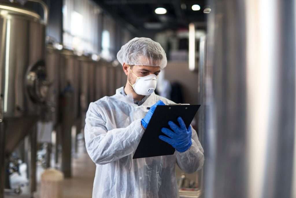 A man in a white coat and gloves holds a clipboard, symbolizing quality management in the pharmaceutical industry.