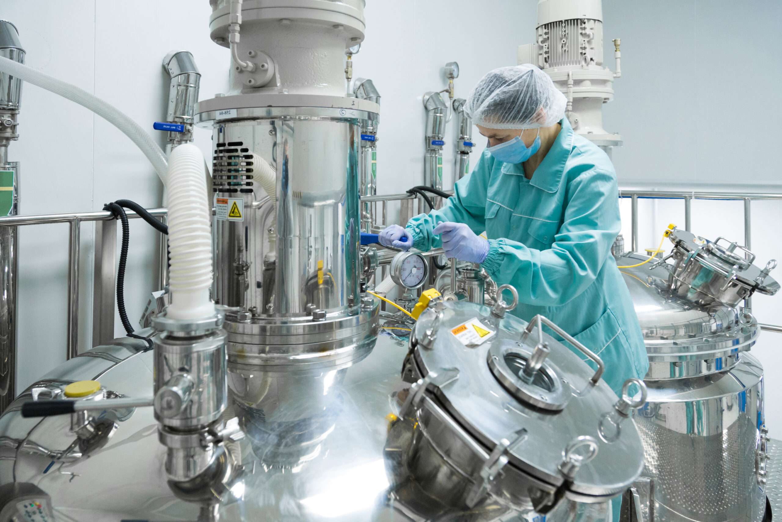 A pharmaceutical factory worker wearing protective clothing and a mask operates stainless steel processing equipment inside a sterile manufacturing environment.